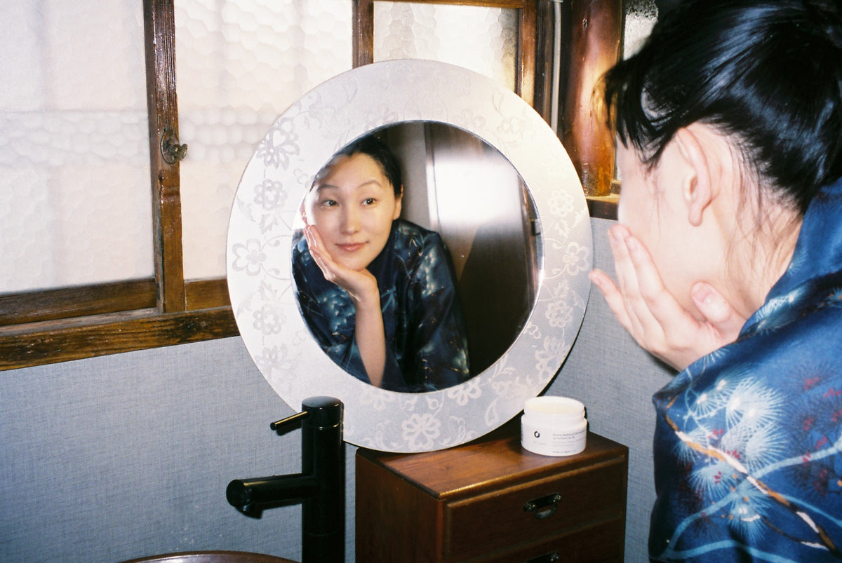 Femme appliquant le baume nettoyant démaquillant Bizen au riz fermenté devant un miroir rond.